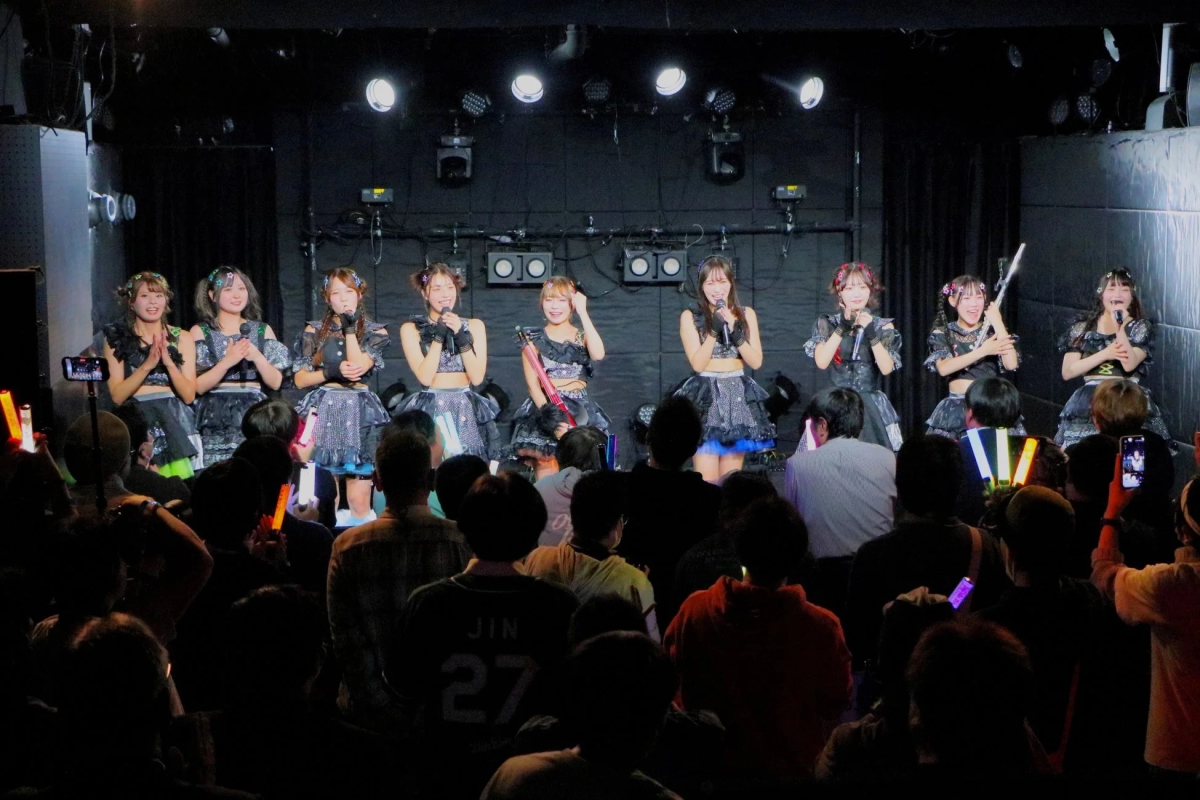 A vibrant stage shot of the i-Okey idol group performing, with colorful lights and an engaged audience