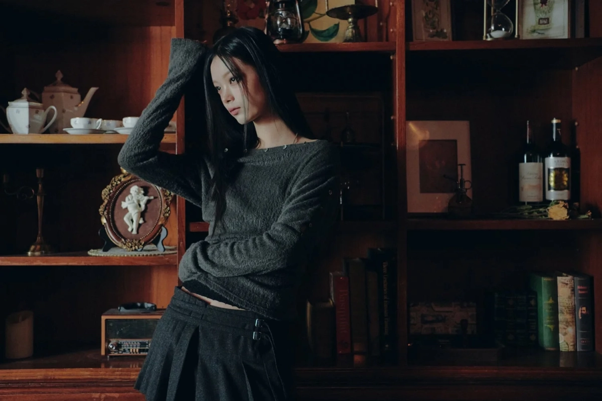 A woman in a grey sweater and pleated skirt sits in a library-like setting, embodying a classic yet modern aesthetic.