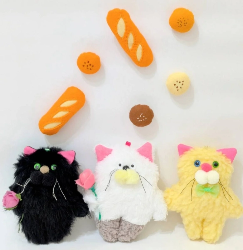 Three fluffy cat plushies in black, white, and yellow, surrounded by felt bread toys