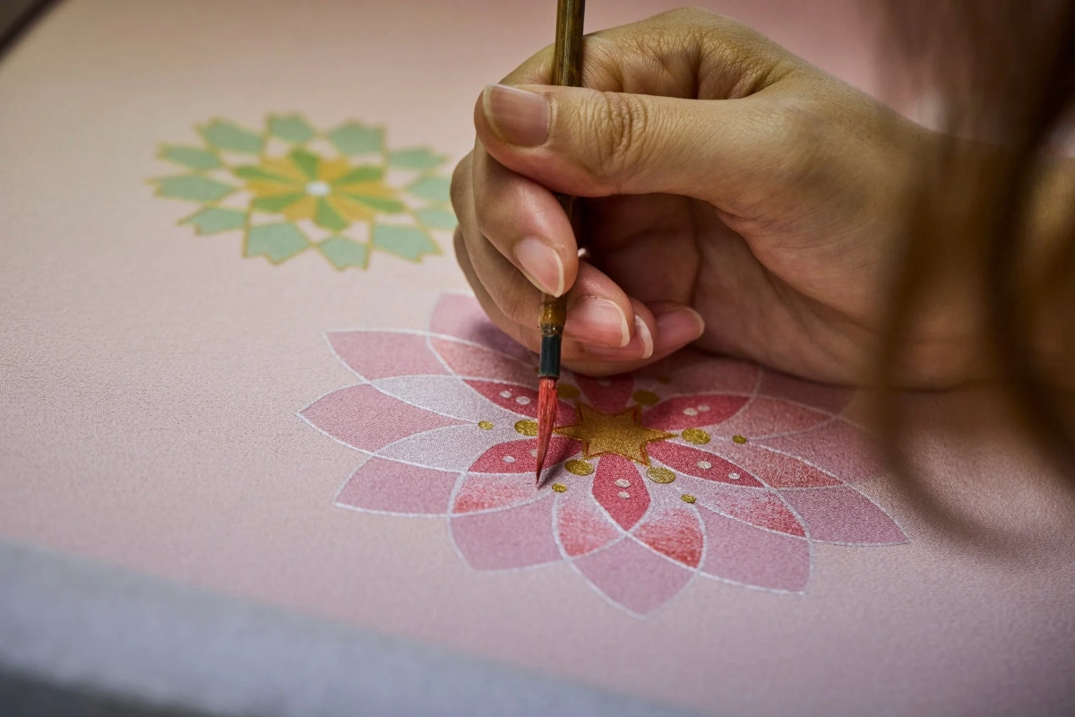 A close-up of a hand applying gold embellishments with a fine brush to a pink fabric with a floral pattern