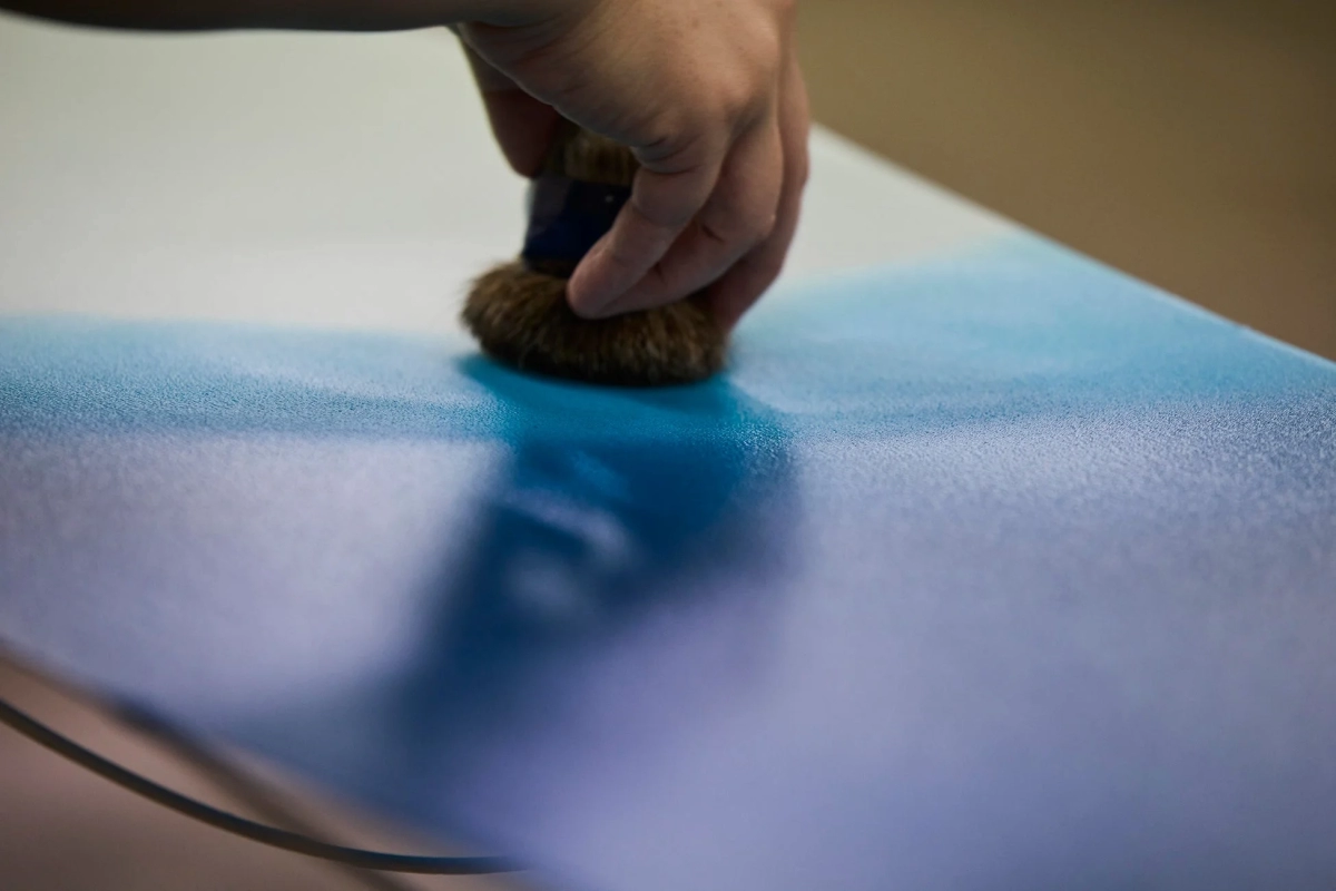 A close-up of a hand using a brush to apply blue dye or paint to a flat surface, creating a delicate gradation