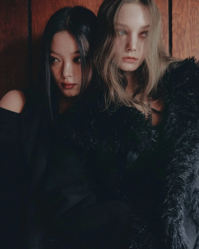 Two women pose in dark clothing, one with black hair looking away, the other with blonde hair facing the camera, creating a mysterious fashion statement.