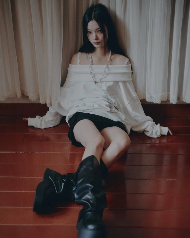 A woman in a white off-shoulder top and black shorts sits on a wooden floor, exuding a cool, casual vibe.