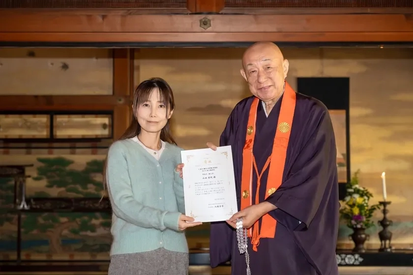 A woman receiving a letter of appreciation from a monk in a traditional setting.