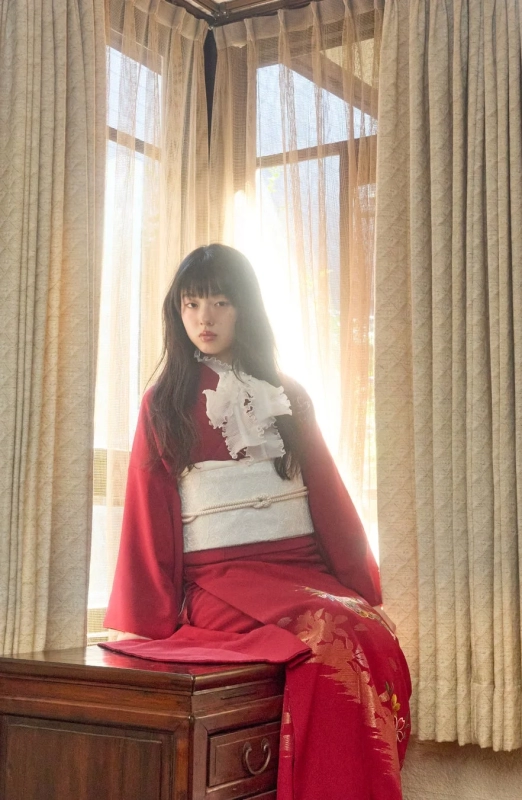 A young woman in a red kimono with white ruffles, sitting by a window