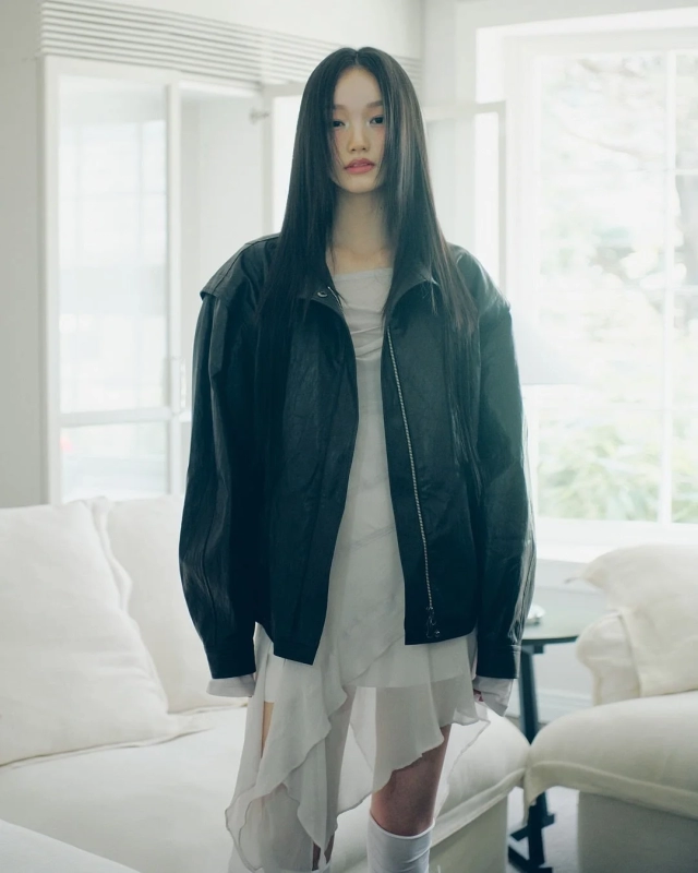 A woman in a black jacket and white ruffled dress stands in a bright room with a white sofa, embodying a stylish and natural aesthetic.