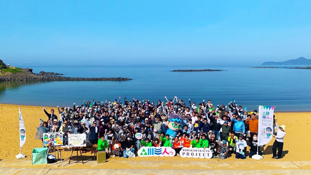 A group of people, including children, on a beach with banners for the "MORIKAWAKAITO PROJECT"