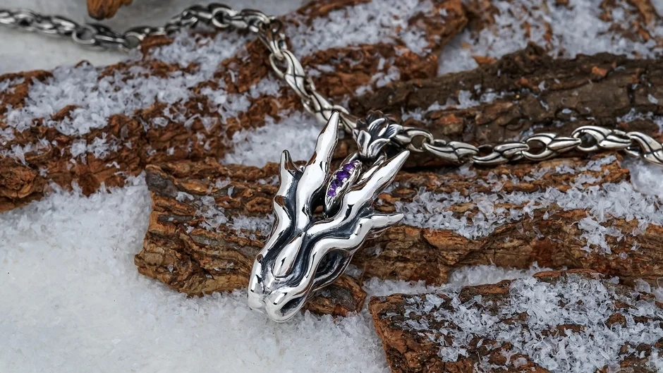 A silver dragon-head pendant adorned with a purple amethyst, displayed on snow and tree bark.