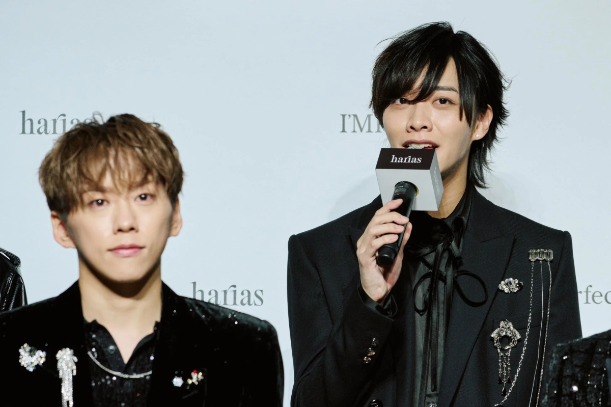 Two IMP. members on stage with HARIAS branding in the background