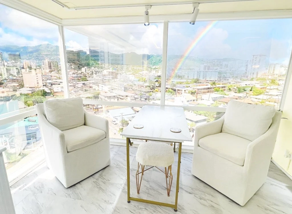 A bright room with a large window overlooking a city and mountains, with a rainbow visible.
