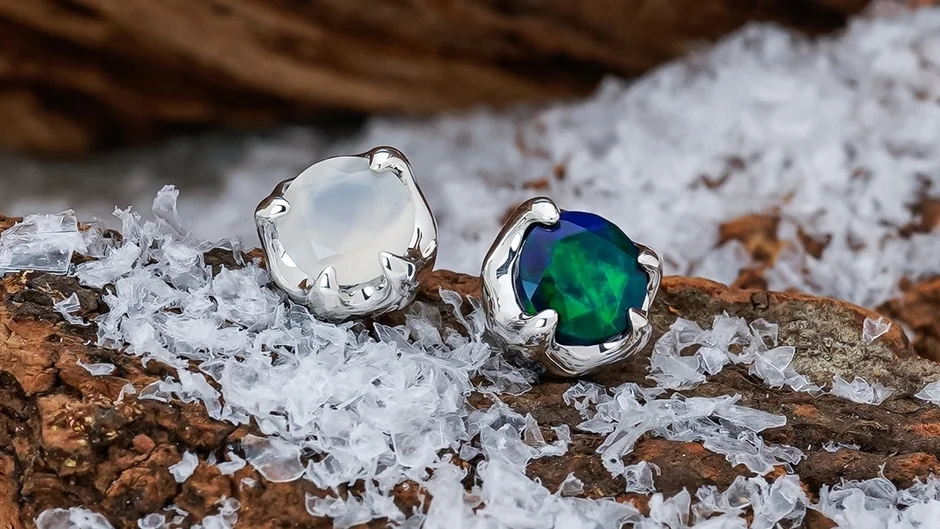 Two exquisite rings or earrings, one with an iridescent opal and the other with a deep green stone, resting amidst ice fragments on a wooden surface.