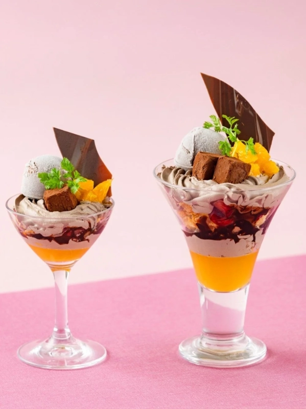Two elegant parfaits in glasses, layered with chocolate ice cream, orange jelly, and chocolate cream, garnished with chocolate decorations and green herbs.