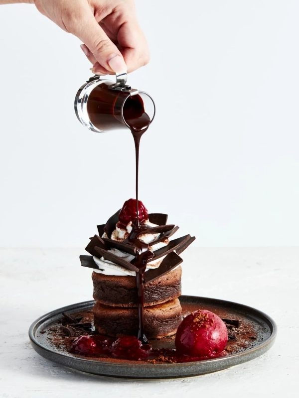 Stacked chocolate cakes drizzled with chocolate sauce, adorned with whipped cream, chocolate chips, and red berries, served with berry ice cream and sauce.