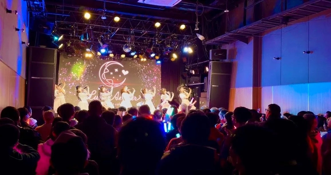 An idol group performs on a stage with colorful lighting, while a large audience waves glow sticks in a lively concert venue.