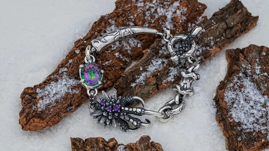 Intricate silver bracelet with mystic topaz and amethyst in a claw-like design, placed on snow-covered tree bark.