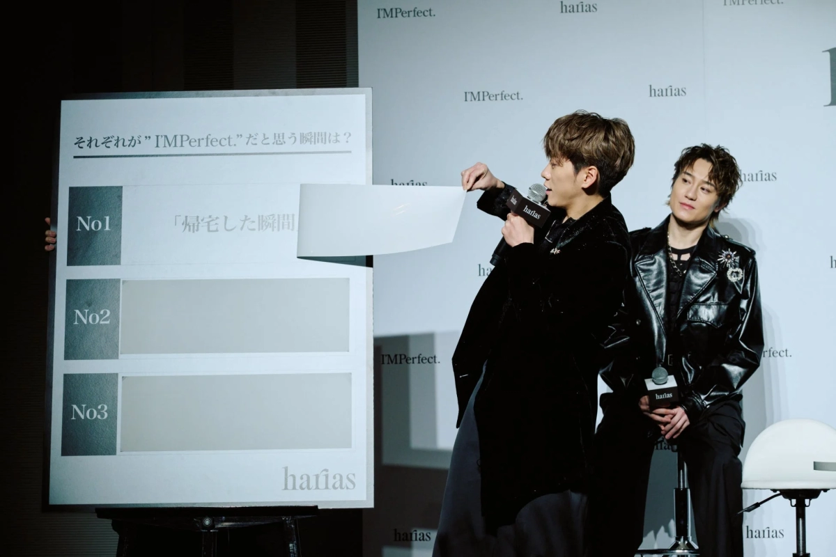 Two IMP. members presenting on stage with an 'I'MPerfect.' panel