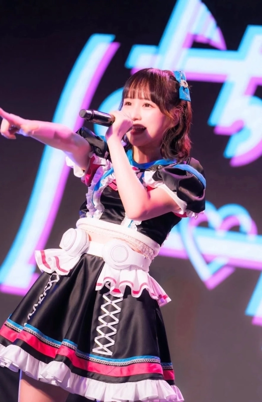 A female idol in colorful stage costume sings with a microphone, pointing with her left hand. Glowing graphics are in the background.