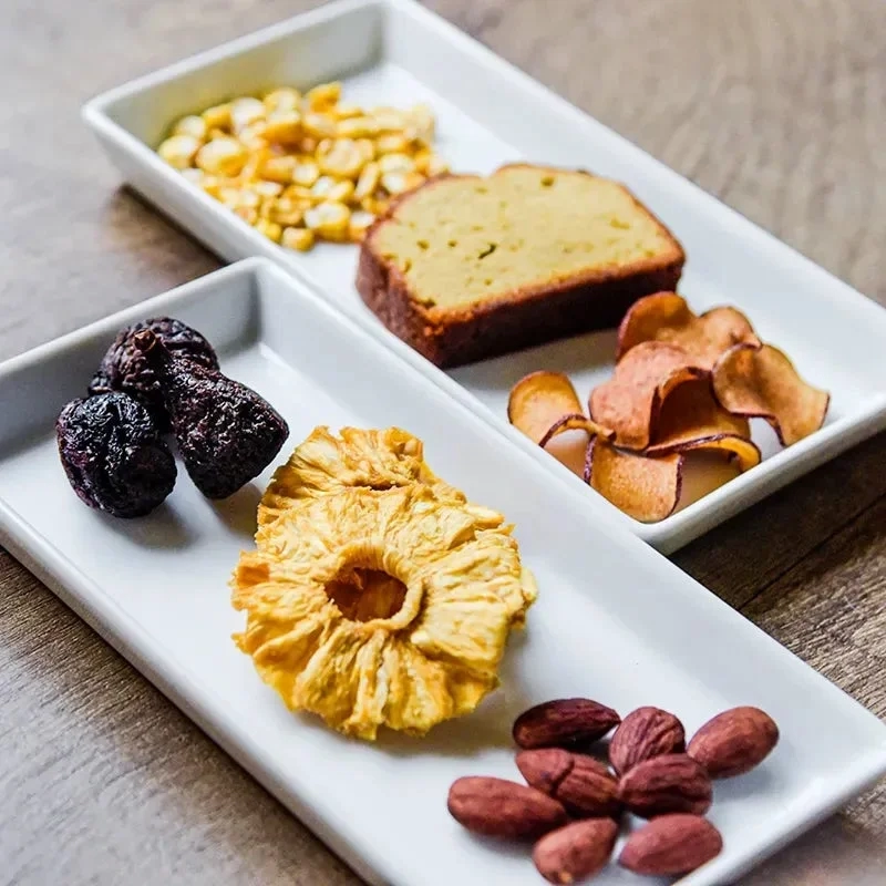 An assortment of healthy snacks including dried pineapple, prunes, almonds, corn, pound cake, and vegetable chips on a white rectangular plate.