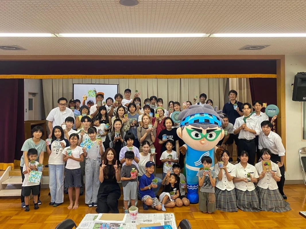 A group of adults and children holding potted succulents, with a mascot character, in an indoor setting