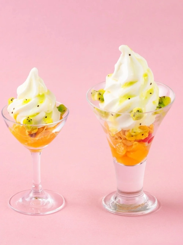 Two vibrant parfaits in glasses, featuring layers of orange fruits, cornflakes, white soft-serve ice cream, and garnished with diced kiwi and green sauce.