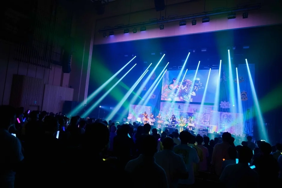 Audience members waving glow sticks at a live concert with performers on a brightly lit stage.