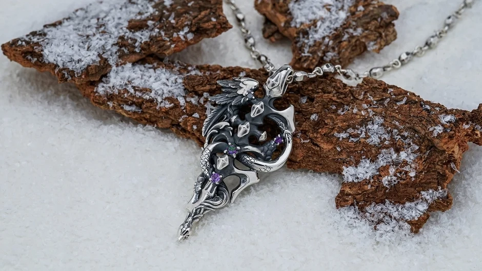 A sophisticated silver necklace pendant featuring a detailed dragon or snake motif with a striking purple gem, nestled on snow-dusted tree bark.