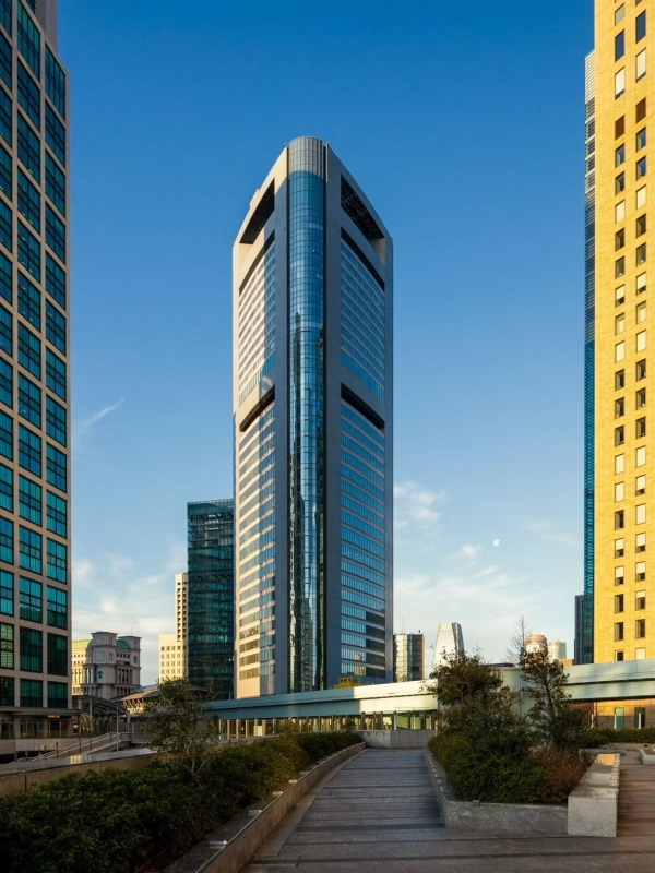 A distinctive modern skyscraper stands under a clear blue sky, surrounded by other urban buildings and green pathways, embodying a contemporary business district.