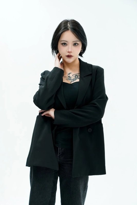 A woman with black hair, wearing a black blazer and top, with visible tattoos and a moon necklace.