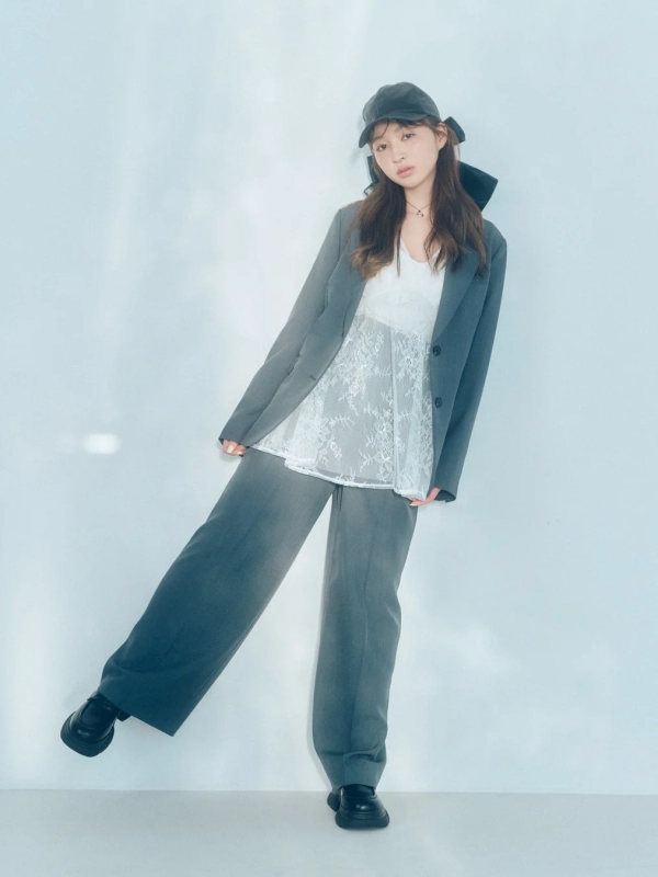 A woman in a grey blazer and wide pants with a white lace top and cap