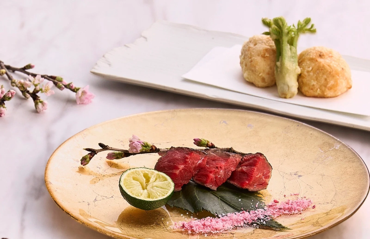 A beautifully plated meat dish with a cherry blossom branch and lime, alongside tempura, arranged on a golden plate for a refined Japanese meal.