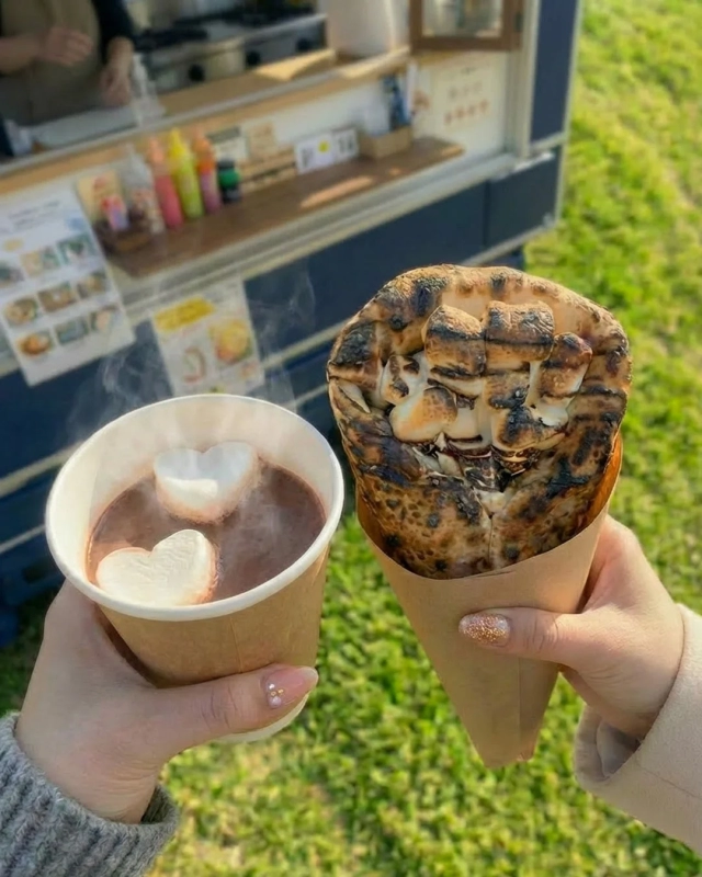 A hot chocolate with heart-shaped marshmallows and a crepe filled with toasted marshmallows, held outdoors.
