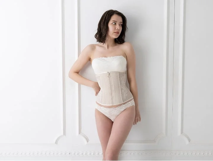 Woman in a strapless bra, corset, and briefs against a white wall
