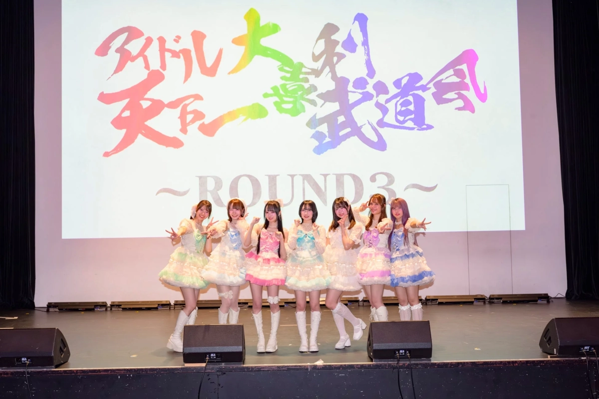 Seven idols in colorful outfits with Budokai screen