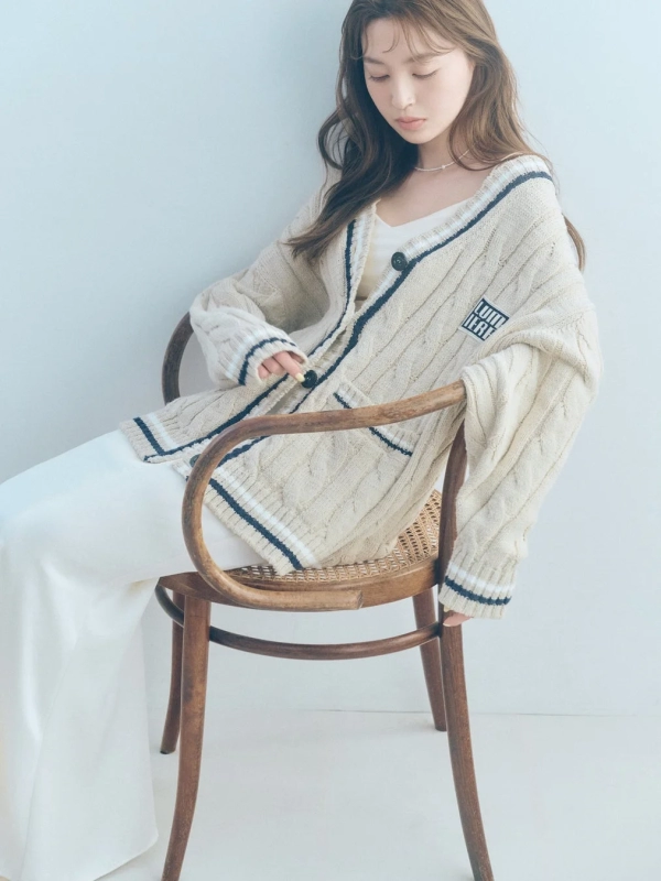 A woman in a beige cable knit cardigan and white pants, sitting on a wooden chair