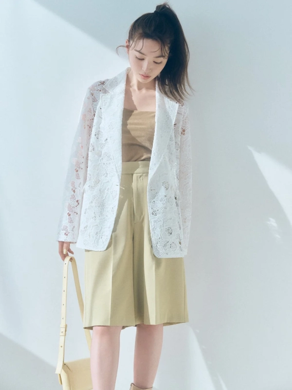 A woman in a white lace jacket and beige shorts, holding a shoulder bag