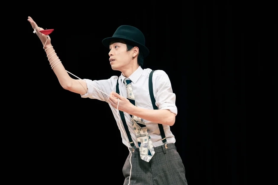 A male performer in a hat and suspenders skillfully juggling a diabolo against a dark background