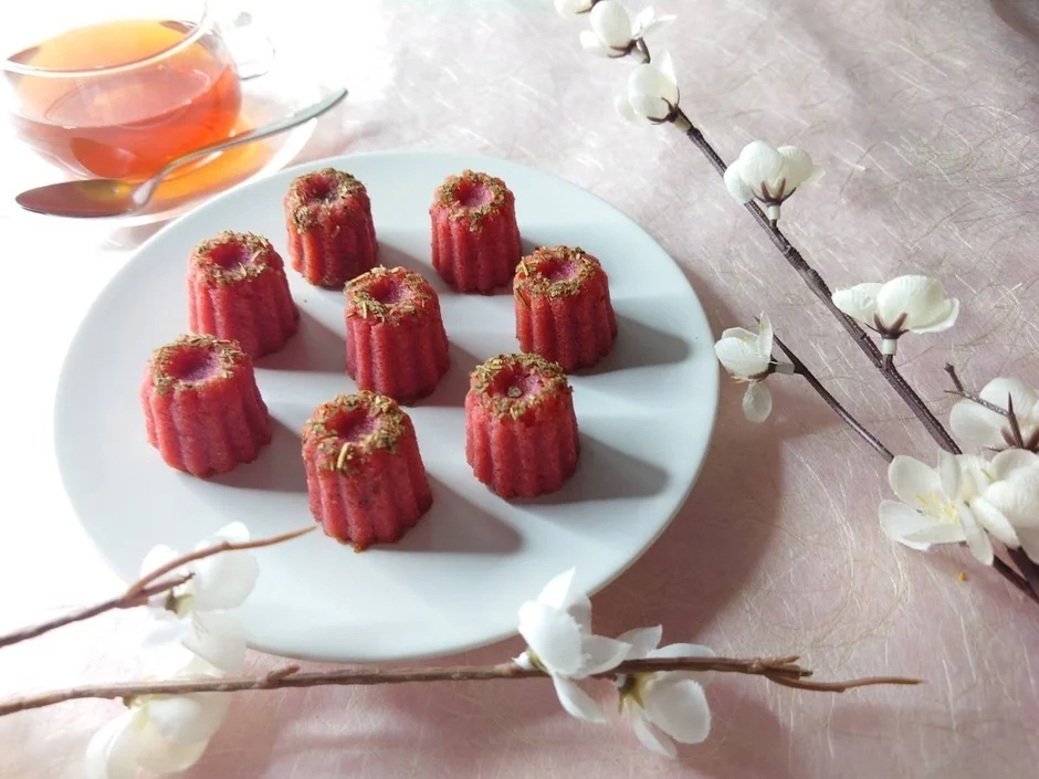 A refined collection of red canelé pastries arranged on a white plate, with a warm cup of tea in the background, suggesting an elegant tea time.