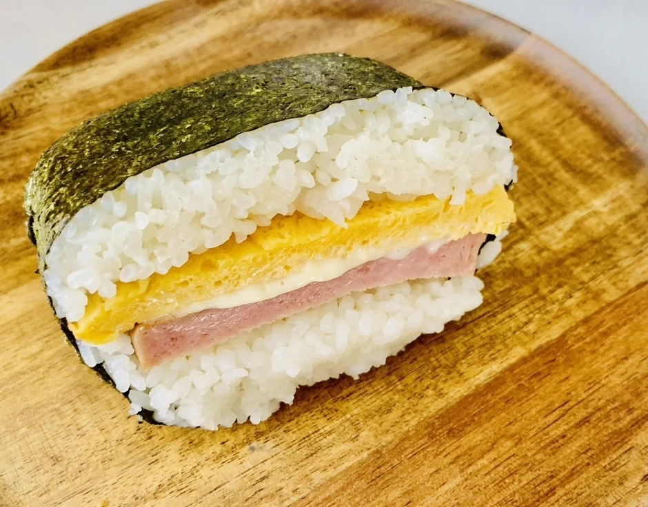 A Spam Onigirazu with a thick omelet, wrapped in nori, on a wooden plate