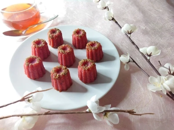 Red canelé-shaped financiers on a white plate with a cup of tea and white flowers