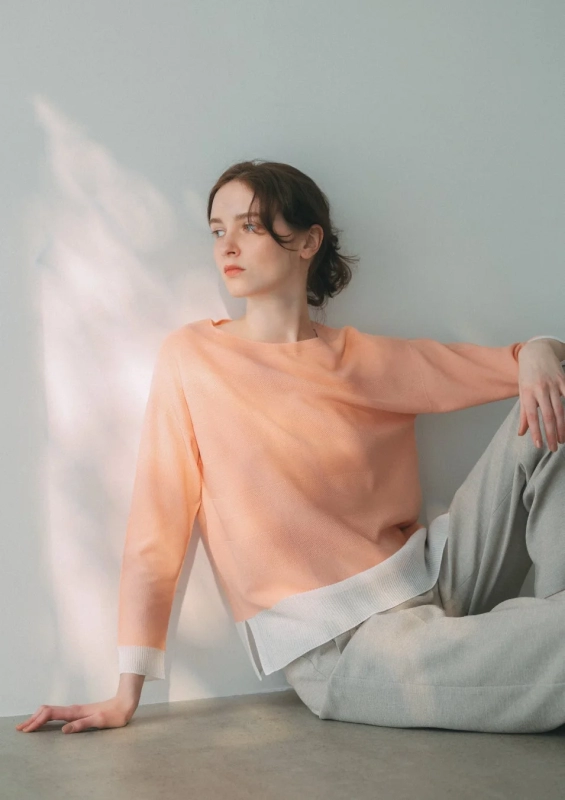A woman in a peach-colored sweater and light gray pants, sitting relaxed against a white wall, embodying the brand's comfortable yet stylish ethos.