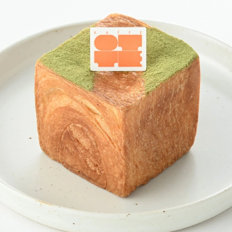 A cube-shaped Danish pastry dusted with matcha powder, served on a white plate.