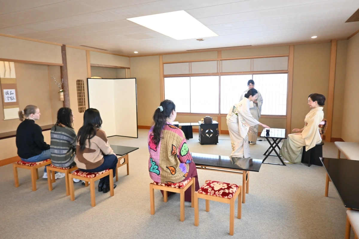 A tea master in a kimono demonstrating the tea ceremony to participants in a Japanese room.