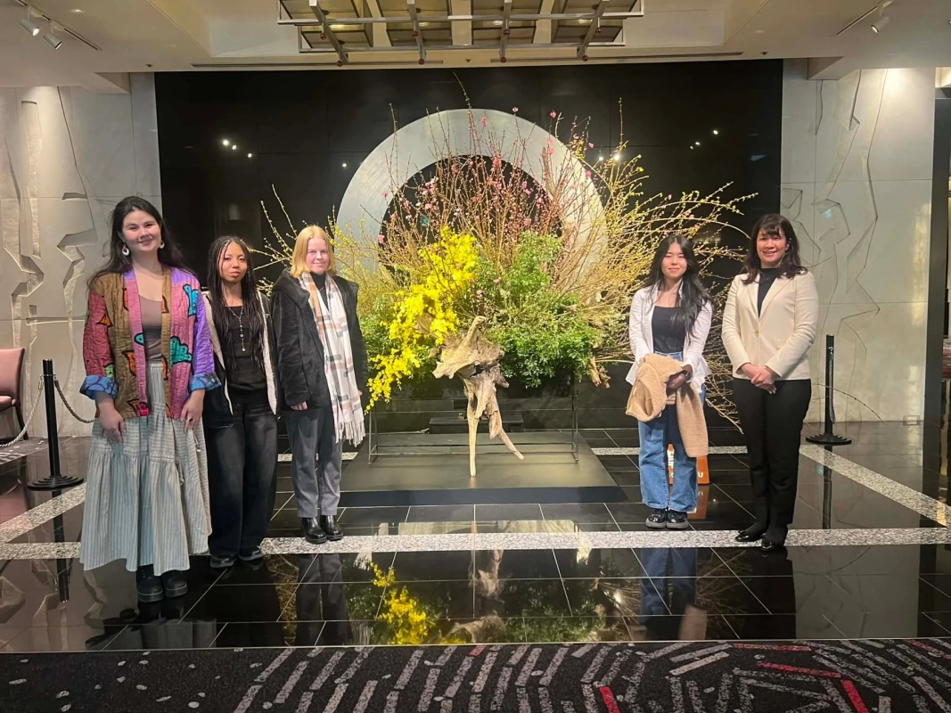 A group of women smiling in a modern lobby, posing in front of a large, vibrant Ikebana arrangement.