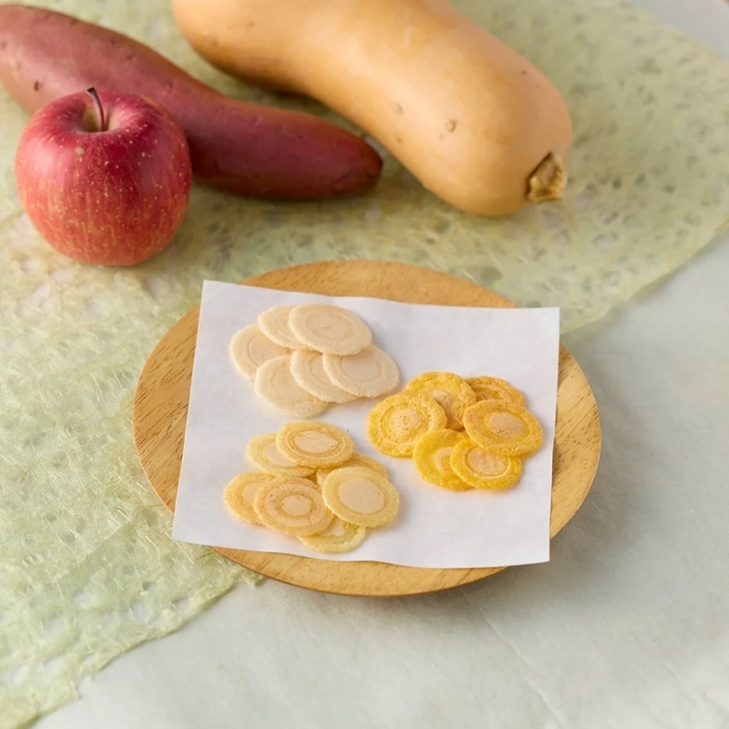 A wooden plate holds various circular snack chips, with raw ingredients like sweet potato, pumpkin, and apple in the background, emphasizing natural ingredients.
