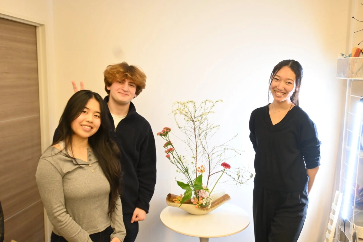Three smiling young people posing indoors with a beautiful Ikebana arrangement on a white table.