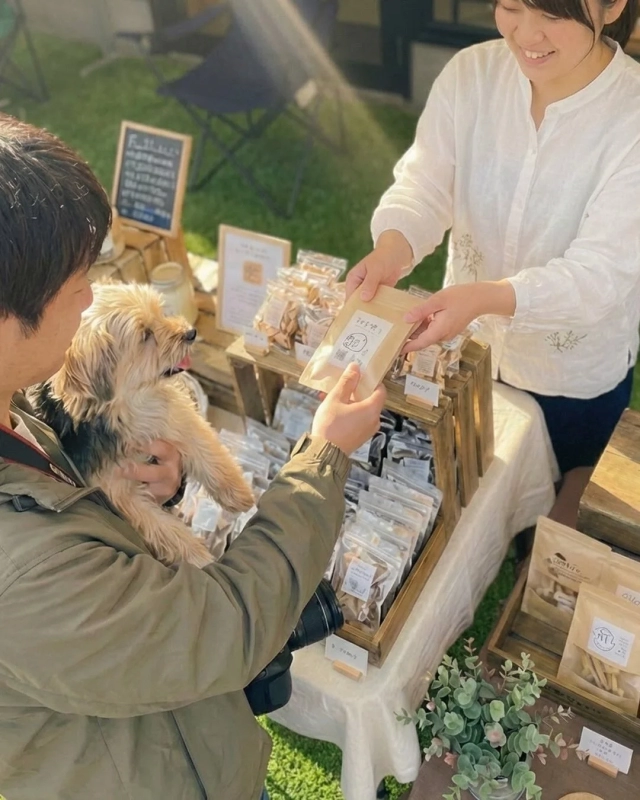 A smiling vendor interacts with a person holding a dog, offering handmade treats at an outdoor market.
