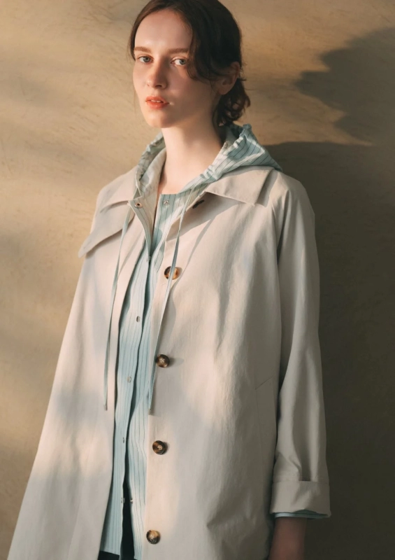 A woman wearing a beige trench coat over a striped shirt, with a relaxed and natural expression.