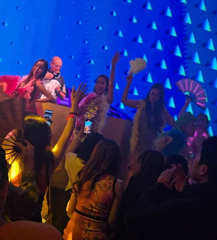 A vibrant club scene with a DJ, dancers wearing feathers, and a crowd holding up smartphones.