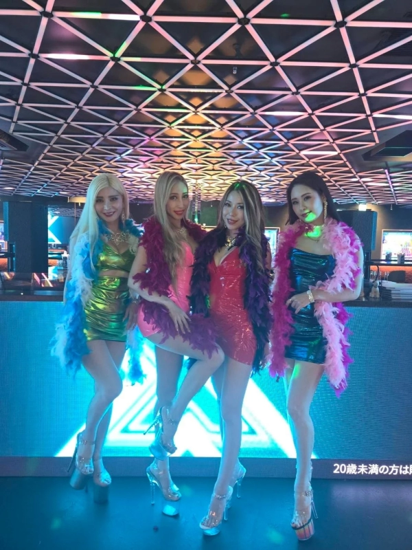 A group of four women in glamorous bodycon dresses and high heels pose in a vibrant, neon-lit club setting.