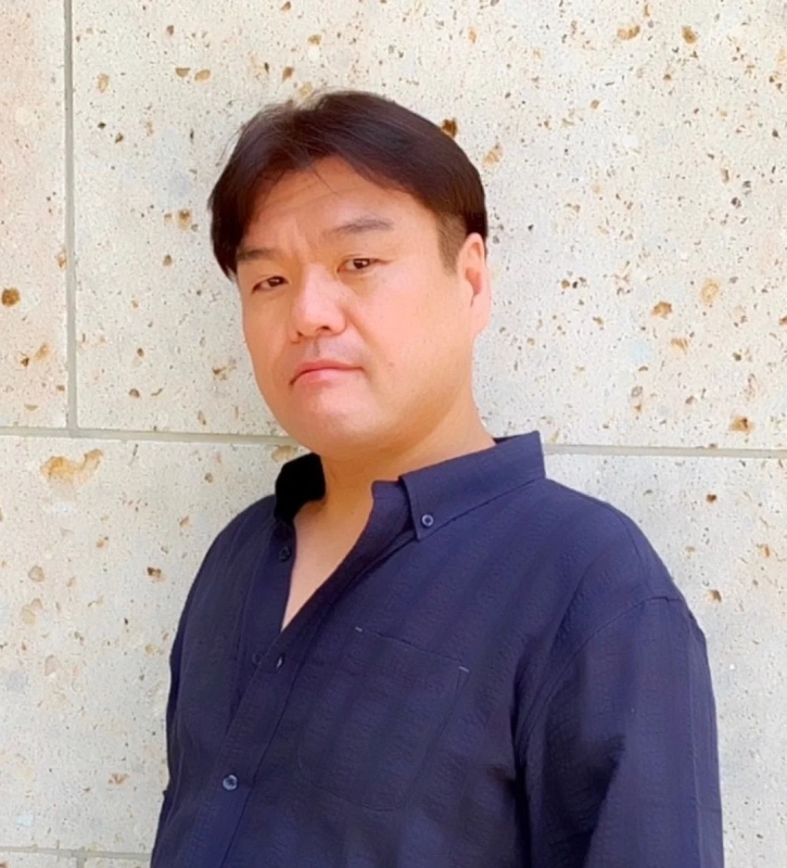 A portrait of an Asian man in a dark blue shirt, looking seriously at the camera against a light-colored wall.